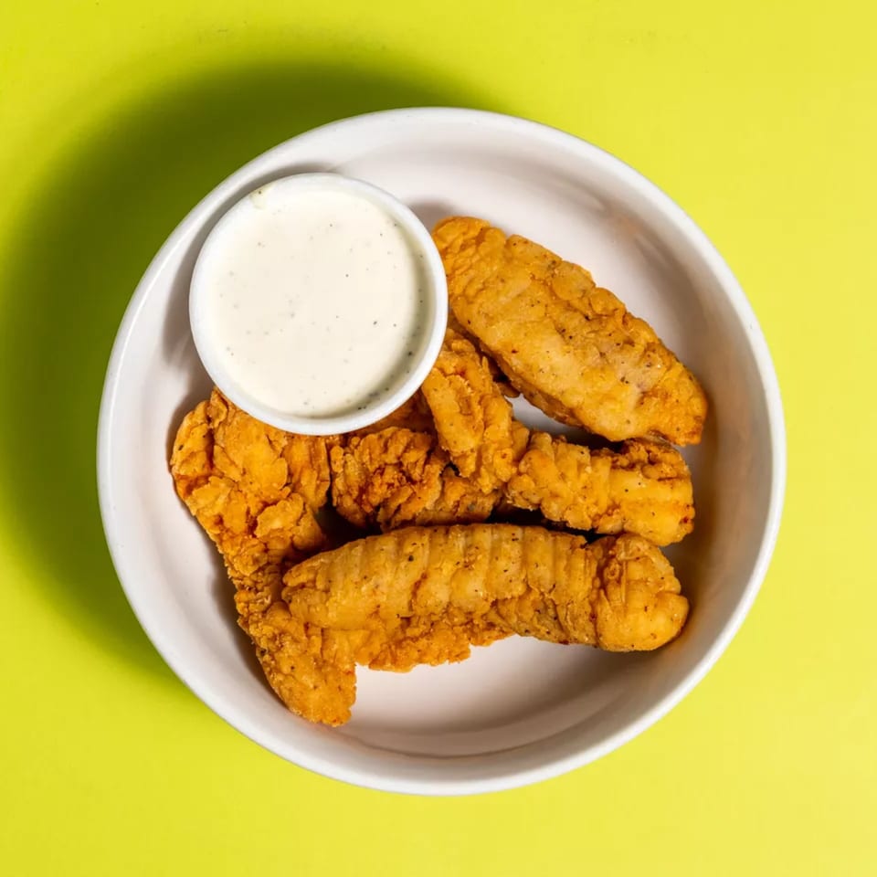 Crispy golden chicken tenders with a side of creamy ranch dipping sauce in a white bowl.
